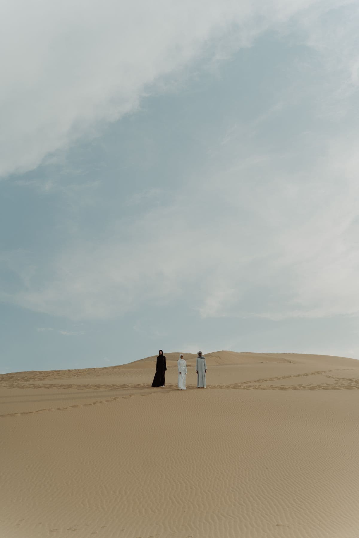 Three women in black and white abayas and hijabs on distant desert dunes under a wide pale sky