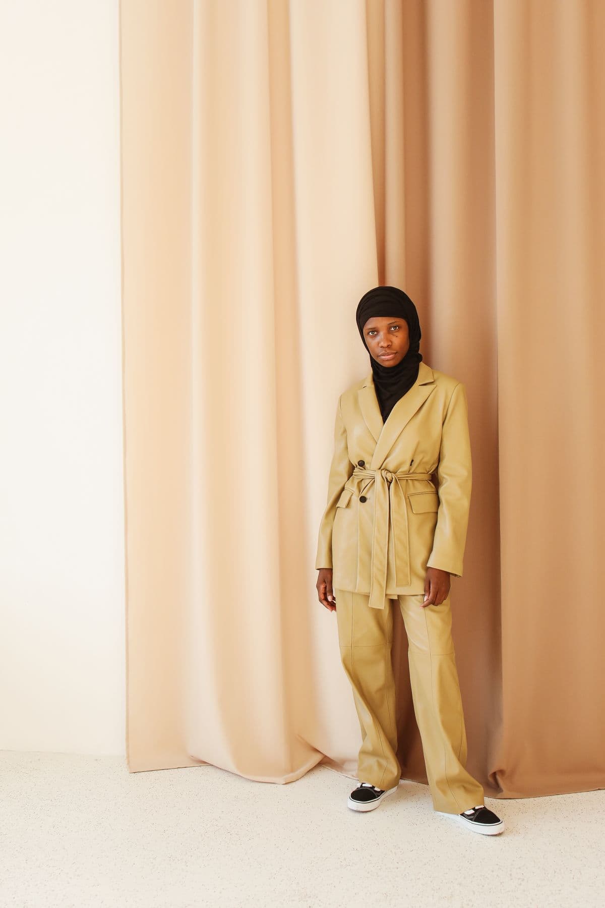 Woman in a hijab and camel belted suit standing against peach curtains in a bright minimal room
